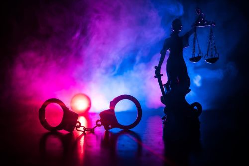 Legal law concept. Silhouette of handcuffs with The Statue of Justice on backside with the flashing red and blue police lights at foggy background. Selective focus